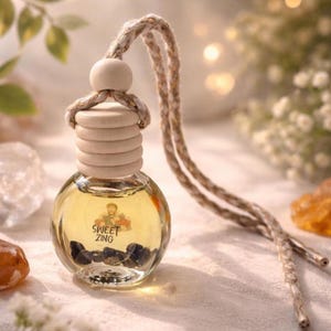May include: A small, round glass bottle filled with a yellow liquid and dark stones. The bottle has a wooden cap and a braided rope for hanging. The text "SWEET ZING" is visible on the bottle. The overall theme is aromatherapy.