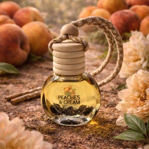 May include: A clear glass bottle with a wooden cap and a rope hanger. The bottle contains a yellow liquid and the words "PEACHES & CREAM" are printed on the front. Peaches and flowers are in the background.