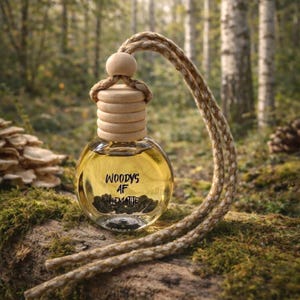 May include: A small, round glass bottle with a wooden cap and a rope hanging from it. The bottle contains a yellow liquid and dark stones. The text "WOODYS AF HEMATITE" is printed on the bottle. The bottle is sitting on a mossy log in a forest setting.