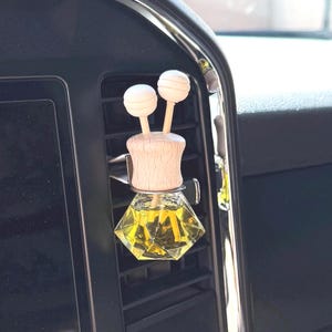May include: Car air freshener with a wooden top and two white ball accents. The clear, geometric-shaped glass bottle contains yellow liquid. The air freshener is clipped to a car vent.