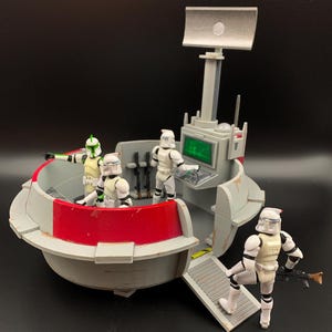 May include: A grey plastic Star Wars command station playset with a red accent panel, a tall radar dish, and a green computer screen. Four white clone trooper action figures are positioned inside and outside the structure, with one holding a rifle.