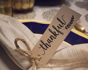 Puede incluir: Una servilleta blanca con rayas doradas está atada con hilo y una etiqueta que dice "thankful" en cursiva. La etiqueta tiene un pequeño diseño de hojas. La servilleta está sobre un plato blanco con un borde azul y dorado.
