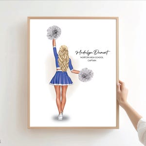 Könnte beinhalten: Ein gerahmter Druck einer blonden Cheerleaderin von hinten, die eine blau-weiße Uniform trägt und zwei silberne Pompons hält. Der Text lautet Madalyn Dumont, Norton High School, Captain. Die Illustration ist vor einem weißen Hintergrund.