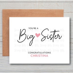 May include: A white card with black text that reads "You're a Big Sister" with a pink heart between the words. The name Christina is written in pink below the text.