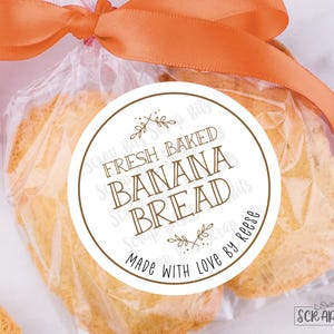 May include: A white round sticker with brown text that reads "Fresh Baked Banana Bread Made with Love by Reese". The sticker is on a clear plastic bag with two pieces of banana bread inside.