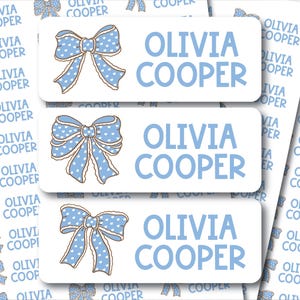 May include: White rectangular labels with the name "OLIVIA COOPER" in light blue. Each label features a light blue bow with white polka dots. The labels are designed for personal use, such as for labeling gifts or belongings.