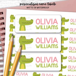 May include: Rectangular white name labels featuring a green cartoon alligator illustration and the text Olivia Williams in pink and green. The labels are displayed on a sheet with multiple repetitions of the design, alongside two yellow pencils.