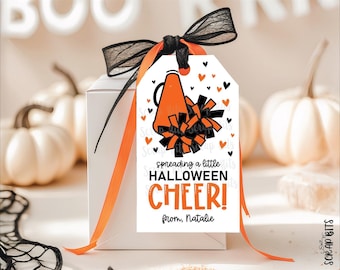 May include: A rectangular white gift tag featuring an orange megaphone and black and orange pom-poms. The text reads, "spreading a little HALLOWEEN CHEER! from, Natalie." The tag is tied with a black and orange ribbon to a white box.