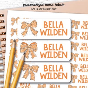 May include: A sheet of rectangular personalized name labels featuring the text BELLA WILDEN in orange font next to an orange polka dot bow illustration. Two yellow pencils are placed diagonally across the labels on a spiral notebook.