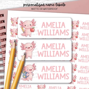Peut inclure: Une feuille d'étiquettes nominatives rectangulaires personnalisées avec un personnage de dessin animé axolotl rose. Chaque étiquette affiche le nom AMELIA WILLIAMS en texte rose. L'axolotl est représenté dans diverses poses, tenant un crayon et une pomme.