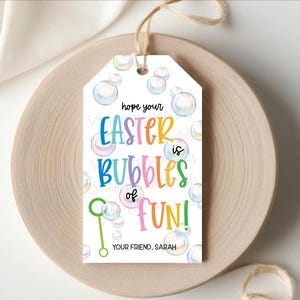 May include: A white gift tag with the text "hope your Easter is Bubbles of FUN!" in colorful letters. The tag features bubble illustrations and a green bubble wand. The tag is tied with twine and rests on a wooden plate.