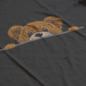 May include: Embroidered brown teddy bear peeking over a silver line on a dark gray fabric. The bear has a fluffy texture with black eyes and a black nose. The design is detailed and appears to be stitched.
