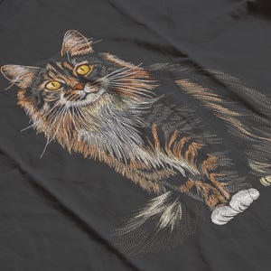 May include: Embroidered design of a long-haired cat with yellow eyes, brown and black fur, and white paws. The cat is lying down on a dark gray surface. The embroidery details the cat's fur and whiskers.