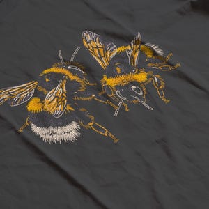May include: Embroidered design of two bumblebees in yellow, black, and white on a dark gray background. The bees are detailed with visible wings, legs, and body segments, showcasing a nature-inspired theme.