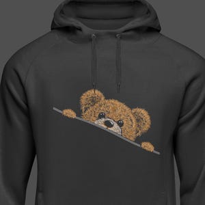 May include: Black hoodie with a brown teddy bear peeking out from the pocket. The teddy bear has a stitched design with a black nose and small, dark eyes. The hoodie has a drawstring and a kangaroo pocket.