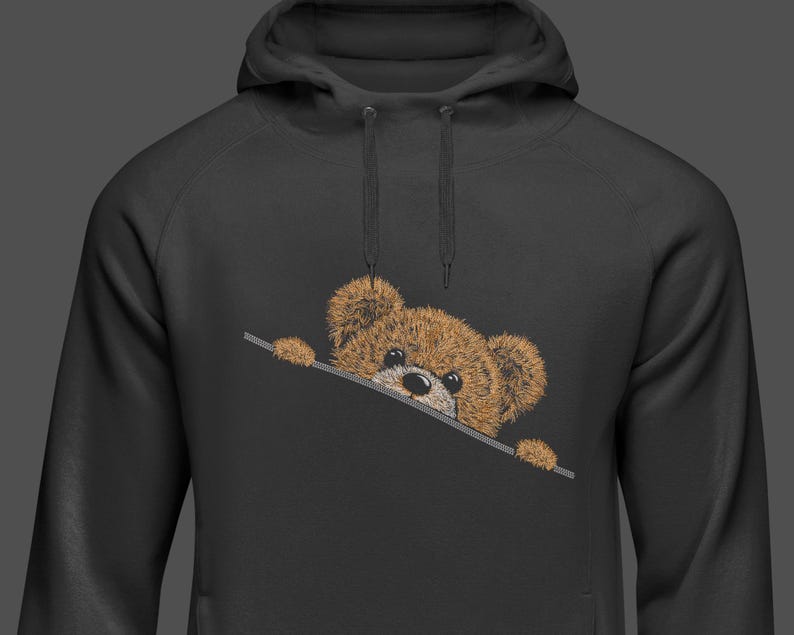 May include: Black hoodie with a brown teddy bear peeking out from the pocket. The teddy bear has a stitched design with a black nose and small, dark eyes. The hoodie has a drawstring and a kangaroo pocket.