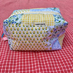 May include: A rectangular fabric pouch featuring a patchwork design of yellow gingham, yellow floral prints, and blue floral patterns. The bag has a top zipper closure and a small white label centred on the front that reads KASHI'S CRAFTS.