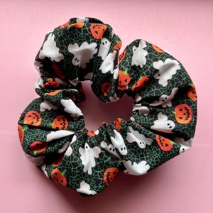 Op de afbeelding: Een Halloween-scrunchie met een zwarte achtergrond, witte geesten en oranje pompoenen. De stof heeft een spinnenwebpatroon. Een haaraccessoire voor het griezelige seizoen.