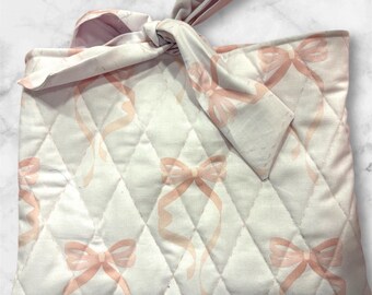 Könnte beinhalten: Eine gesteppte, cremefarbene Stofftasche mit einem sich wiederholenden Muster aus rosa Schleifen und Bändern. Die Tasche ist oben mit einer passenden Stoffschleife gebunden. Die Tasche hat ein Rautenmuster.