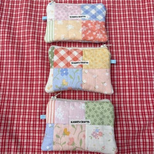 May include: Three rectangular quilted zipper pouches featuring patchwork designs in pastel colors including pink, blue, yellow, and green with floral and gingham patterns. Each pouch has a white fabric label reading KASHI'S CRAFTS centered on the front.