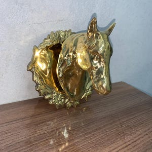 May include: A golden horse head wall decor with a detailed mane and a circular wreath around the opening. The horse head is made of metal and has a shiny finish. The decor is placed on a wooden surface.