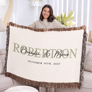 A woman is holding up a large white blanket with the names "Brandy & Maria" and the date "October 15th, 2027" printed on it.
