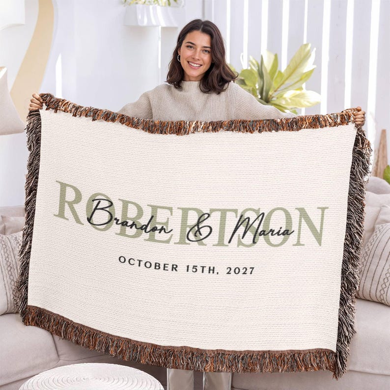 A woman is holding up a large white blanket with the names "Brandy & Maria" and the date "October 15th, 2027" printed on it.