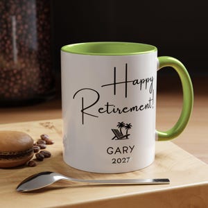 A white coffee mug with a green handle, featuring the text "Happy Retirement! GARY 2027" printed on it. The mug is placed on a wooden surface, with a spoon nearby.