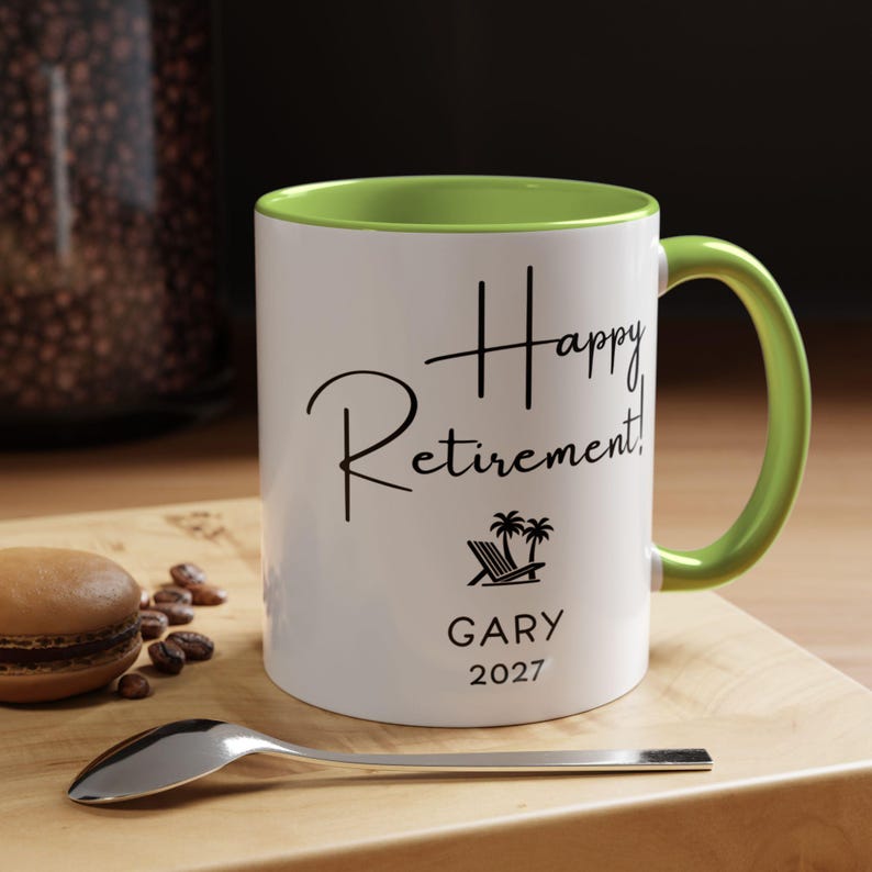 A white coffee mug with a green handle, featuring the text "Happy Retirement! GARY 2027" printed on it. The mug is placed on a wooden surface, with a spoon nearby.
