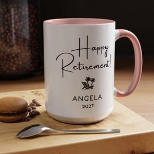 A white ceramic mug with a pink handle, featuring the text "Happy Retirement! Angela 2027" printed on it. The mug is placed on a wooden surface, with a spoon and some coffee beans nearby.
