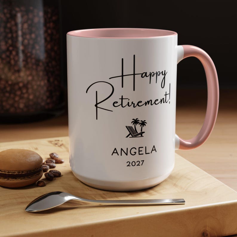 A white ceramic mug with a pink handle, featuring the text "Happy Retirement! Angela 2027" printed on it. The mug is placed on a wooden surface, with a spoon and some coffee beans nearby.
