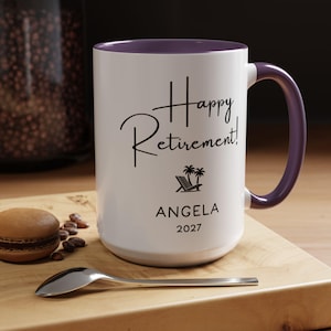 A white ceramic mug with a purple handle, featuring the text "Happy Retirement!" and the name "Angela" along with the year "2027" printed on it. The mug is placed on a wooden surface, with a spoon and some coffee beans nearby.