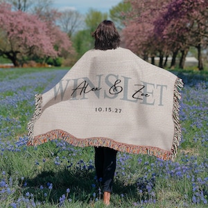 A person standing in a field of flowers, holding a blanket with the names 'Alex & Zoe' and the date '10.15.27' printed on it.