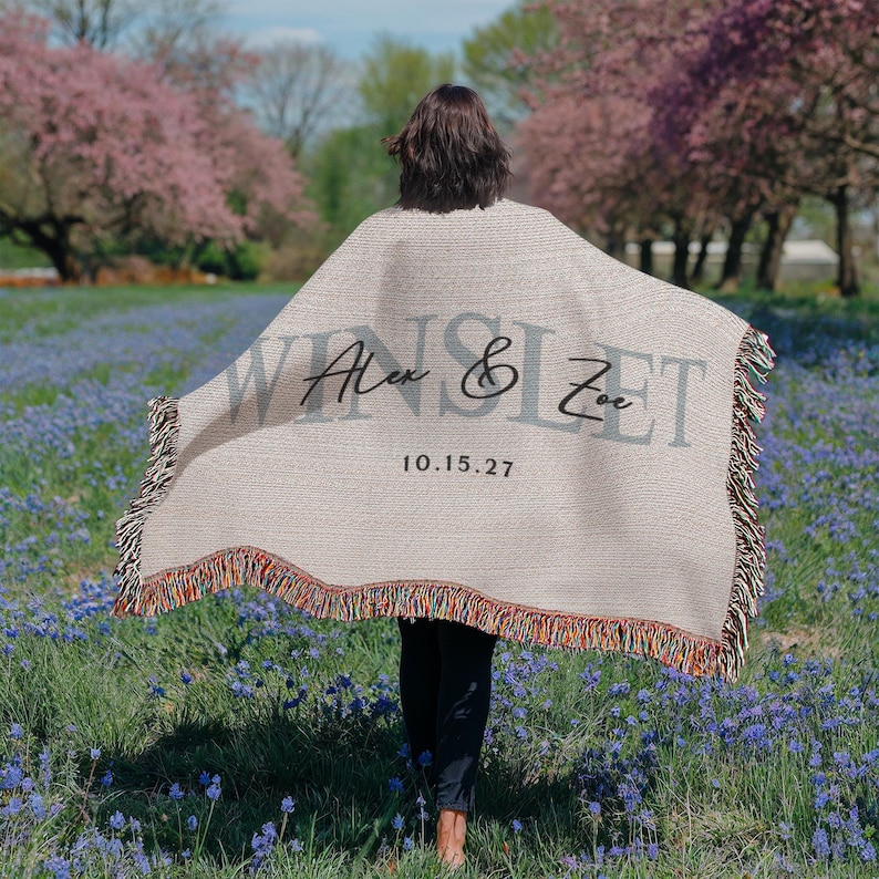 A person standing in a field of flowers, holding a blanket with the names 'Alex & Zoe' and the date '10.15.27' printed on it.