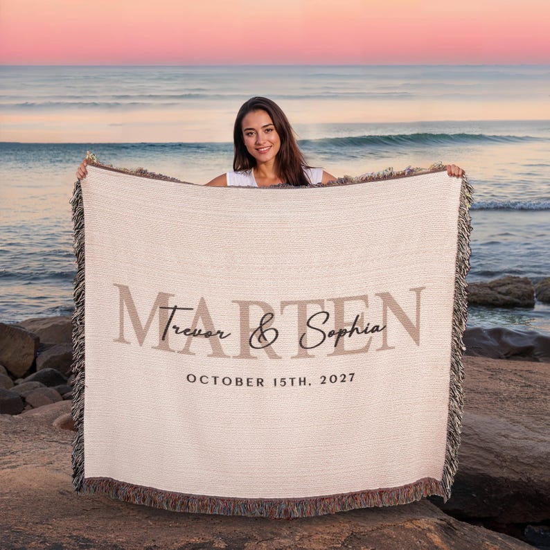A woman is holding a large blanket with the text "Marten" and "Sophia" printed on it, standing on a beach with the ocean in the background.