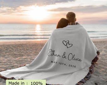 May include: A light gray woven throw blanket with fringed edges, featuring a black printed design of two interlocking hearts above the text Ethan & Olivia and the date MAY 15TH, 2026. The blanket is labeled as 100% Cotton and Made in USA.