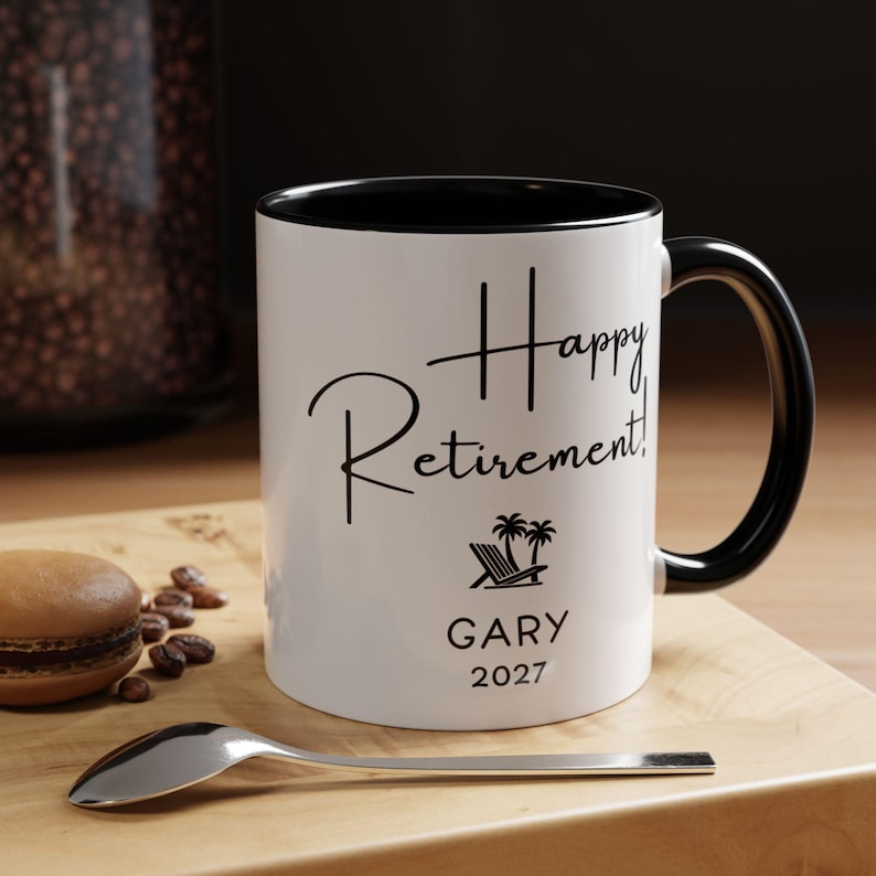 A white coffee mug with black text and a palm tree graphic, sitting on a wooden surface next to a spoon and some coffee beans.