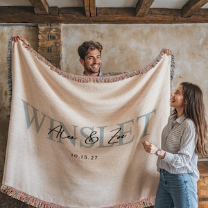 A man and a woman are holding a large beige blanket with the text "WINSET" and the date "10.15.27" printed on it.