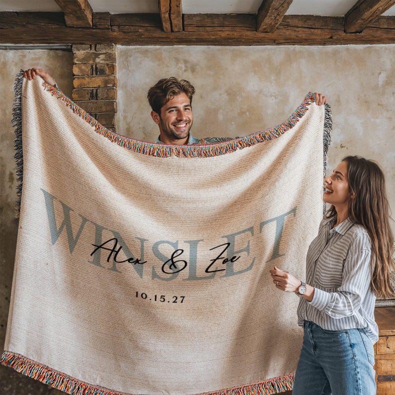 A man and a woman are holding a large beige blanket with the text "WINSET" and the date "10.15.27" printed on it.