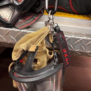 May include: A black and clear respirator mask with red accents, attached to a carabiner. A tan glove and a leather tag with the letters "GKD3" in red are also visible. The items are hanging on a metal surface.