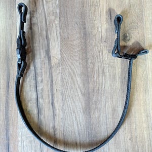 May include: A black adjustable strap featuring a durable plastic side-release buckle and two looped ends. The strap is constructed from a textured material with visible stitching along the edges, designed for securing or carrying items.