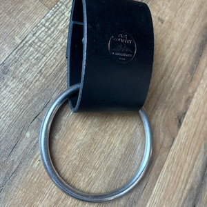 Puede incluir: Un bucle de cinturón de cuero negro con un anillo de metal plateado. El bucle del cinturón tiene el texto "The Country" y "Leather Works" grabado en relieve.