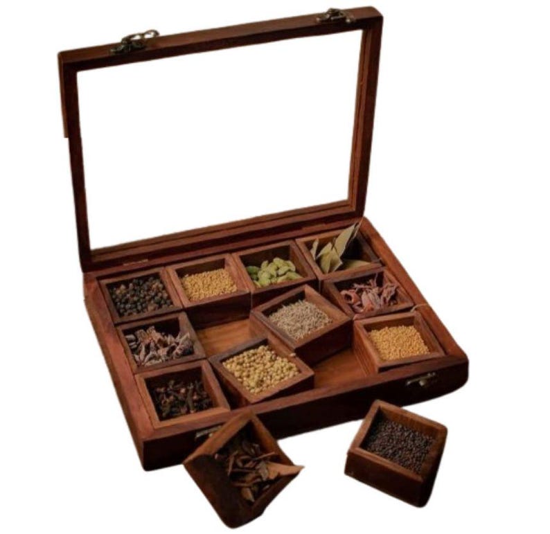 Puede incluir: Una caja de especias de madera con tapa de vidrio, que contiene ocho compartimentos llenos de diferentes especias. La caja est&aacute; hecha de madera marr&oacute;n oscura y tiene una tapa con bisagras. Los compartimentos est&aacute;n dispuestos en forma de cuadr&iacute;cula y est&aacute;n llenos de diferentes especias, incluyendo granos de pimienta negra, clavos, semillas de comino y semillas de cilantro.