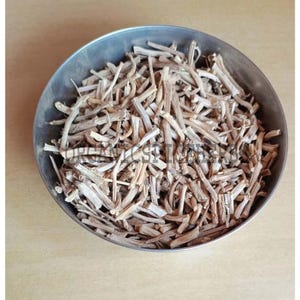 May include: A metal bowl filled with dried, light brown, stick-like pieces. The bowl is round and has a silver color. The sticks are of varying lengths and are densely packed together. The background is a light tan color.