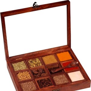 Puede incluir: Una caja de especias de madera con tapa de vidrio, dividida en nueve compartimentos. La caja est&aacute; llena de diversas especias, incluyendo comino, cilantro, c&uacute;rcuma y chile en polvo.
