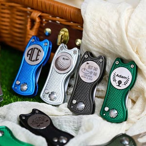 Könnte beinhalten: Eine Sammlung von Golf-Divot-Reparaturwerkzeugen aus Metall in Blau, Silber, Schwarz und Grün. Jedes Werkzeug verfügt über einen strukturierten Griff und eine kreisförmige Mittelplatte, die mit verschiedenen Motiven wie Ace, Ryan, Adam und Best Dad By Par graviert ist.