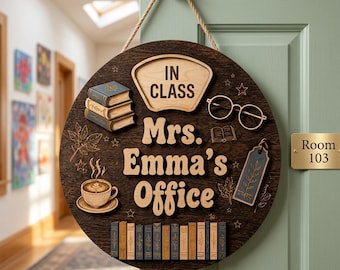 Puede incluir: Un letrero de puerta circular de madera oscura colgado de una cuerda, con el texto "IN CLASS" y "Mrs. Emma's Office" en relieve. El letrero incluye recortes de madera de una pila de tres libros, una taza de café, anteojos y una fila de libros.