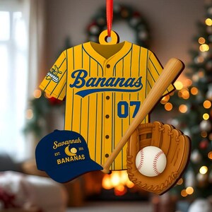 Pode incluir: Um enfeite com tema de beisebol com uma camisa amarela com listras azuis e a palavra "Bananas". Inclui um bast&atilde;o de beisebol, uma luva, uma bola e um bon&eacute; azul que diz "Savannah Bananas". O enfeite &eacute; pendurado em uma fita vermelha.