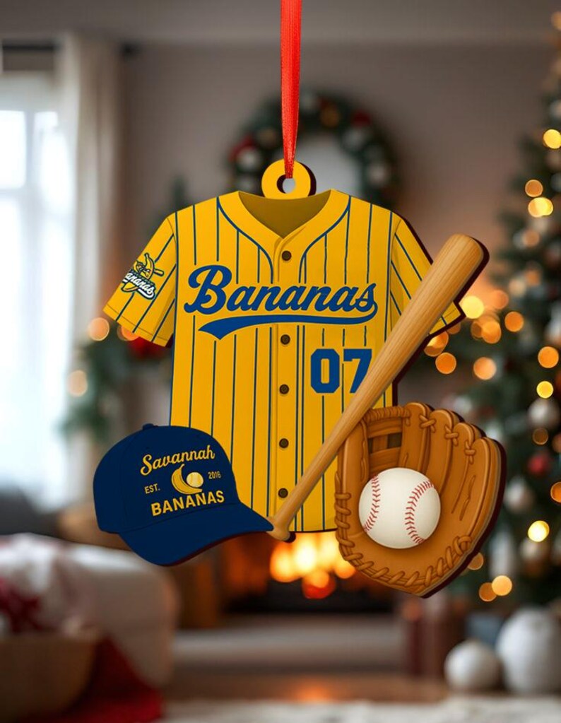 Pode incluir: Um enfeite com tema de beisebol com uma camisa amarela com listras azuis e a palavra "Bananas". Inclui um bast&atilde;o de beisebol, uma luva, uma bola e um bon&eacute; azul que diz "Savannah Bananas". O enfeite &eacute; pendurado em uma fita vermelha.