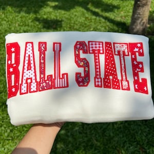 May include: A white sweatshirt featuring the text BALL STATE in large, block-style letters. Each letter is filled with a variety of red and white patterns, including floral prints, polka dots, gingham checks, and star motifs, with a red outline.
